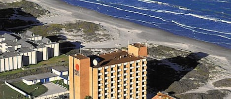 A high-rise hotel with a central tower and multiple balconies, located near a beach with waves crashing onto the shore.
