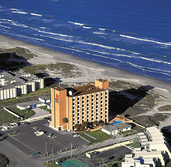 A high-rise hotel with a central tower and multiple balconies, located near a beach with waves crashing onto the shore.