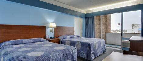 Habitación de hotel con dos camas, un escritorio, una lámpara y un ventanal con vistas a edificios.