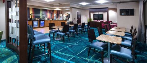 A hotel dining area with blue carpet, tables, chairs, a kitchen area, and a TV on the wall.