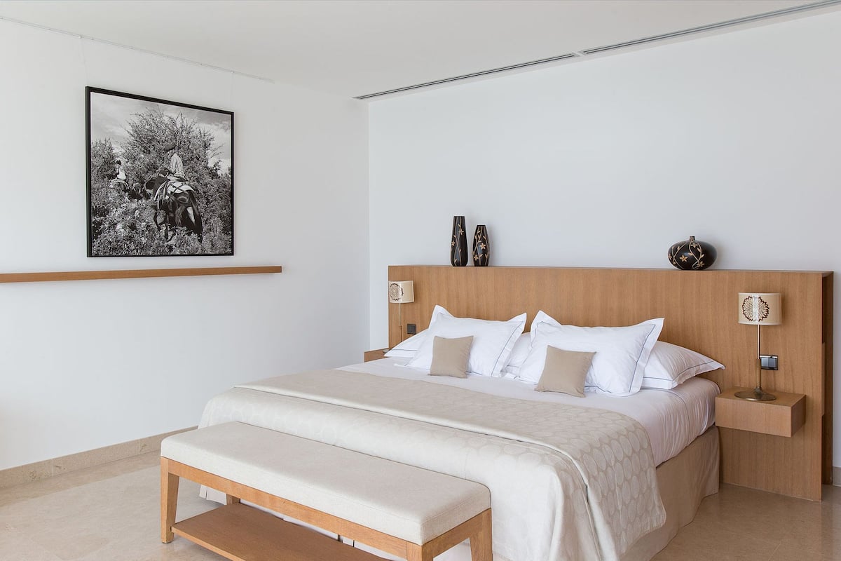 A neatly arranged bedroom with a large bed, a bedside table, a bench, and a wall-mounted shelf with decorative items.