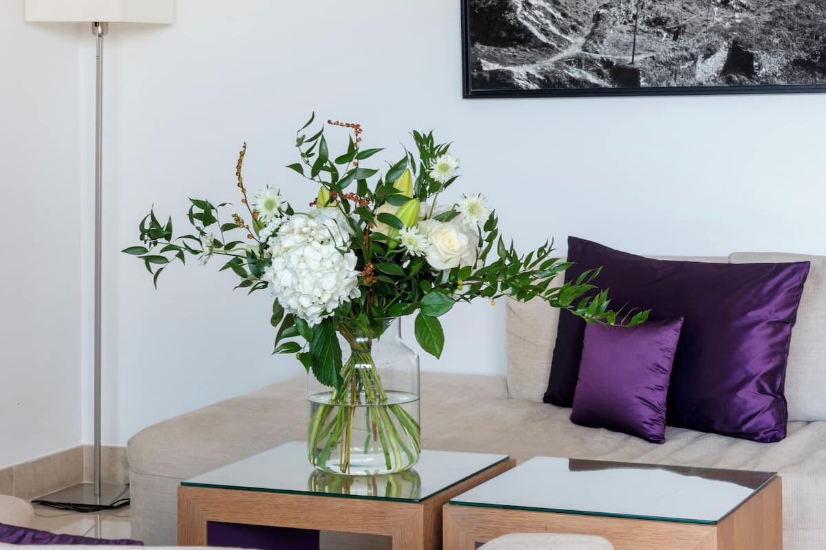 A modern living room with a beige sofa, a glass coffee table, and a vase of flowers. A black and white photograph of a castle is on the wall.