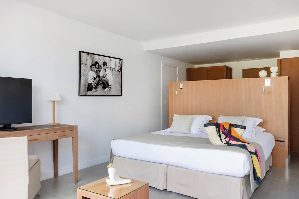 A modern hotel room with a bed, a desk with a lamp, a television, and a framed picture on the wall.