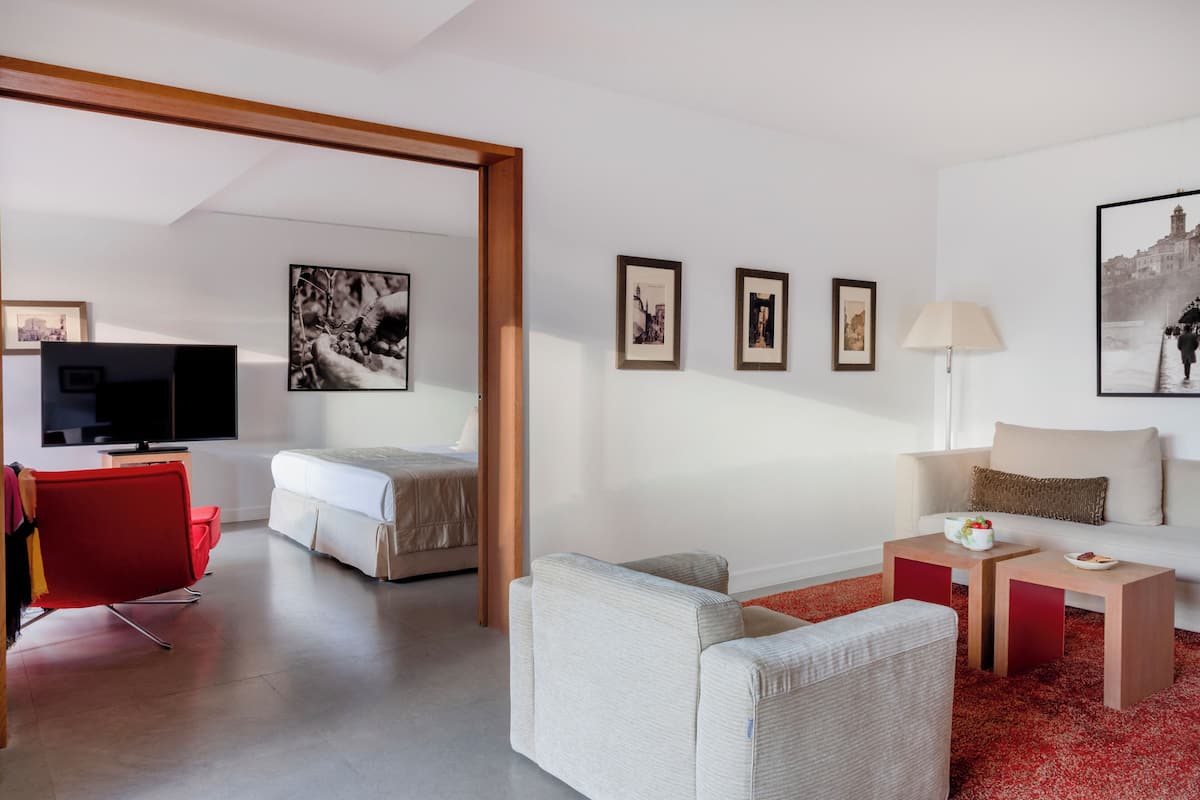 A modern hotel room with a bed, a TV, a red chair, a sofa, and a small table with a lamp.