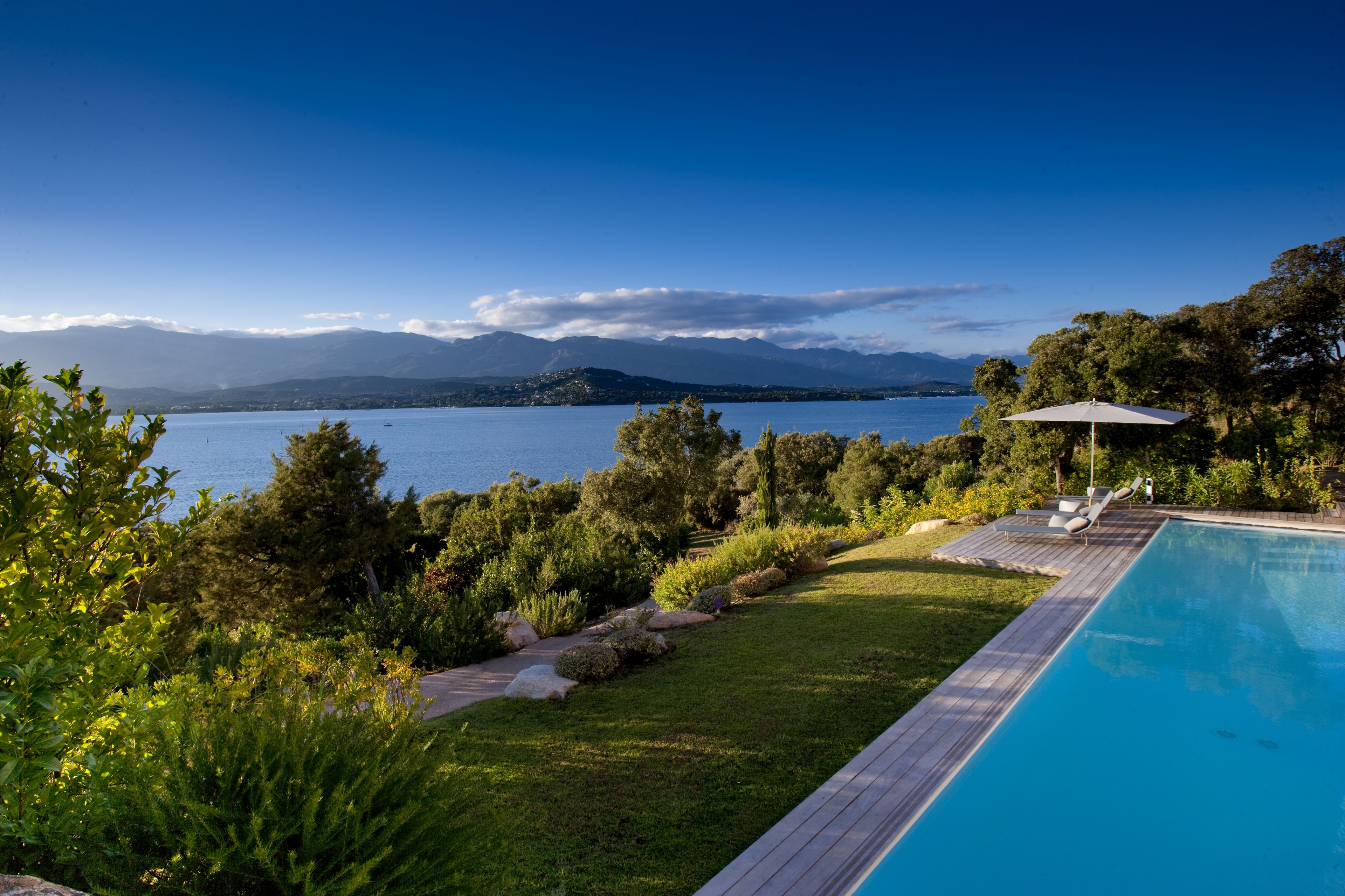 Una piscina con bordo in legno, circondata da vegetazione rigogliosa e vista su un lago con montagne sullo sfondo.