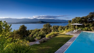 A swimming pool with a wooden deck, surrounded by lush greenery and a view of a lake with mountains in the background.