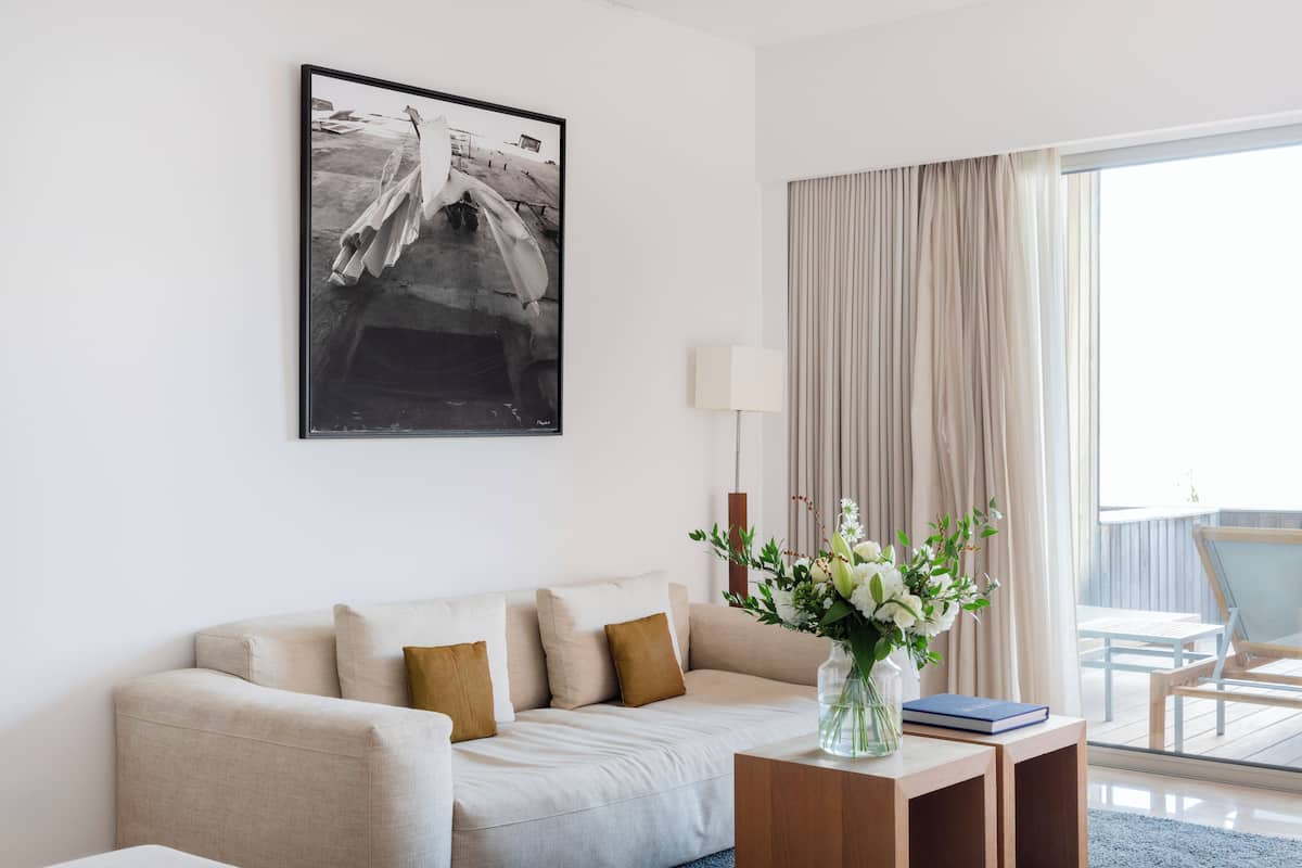 A modern living room with a sofa, a side table with a vase and book, a framed black and white photograph, and a view of the outdoors through a large window.