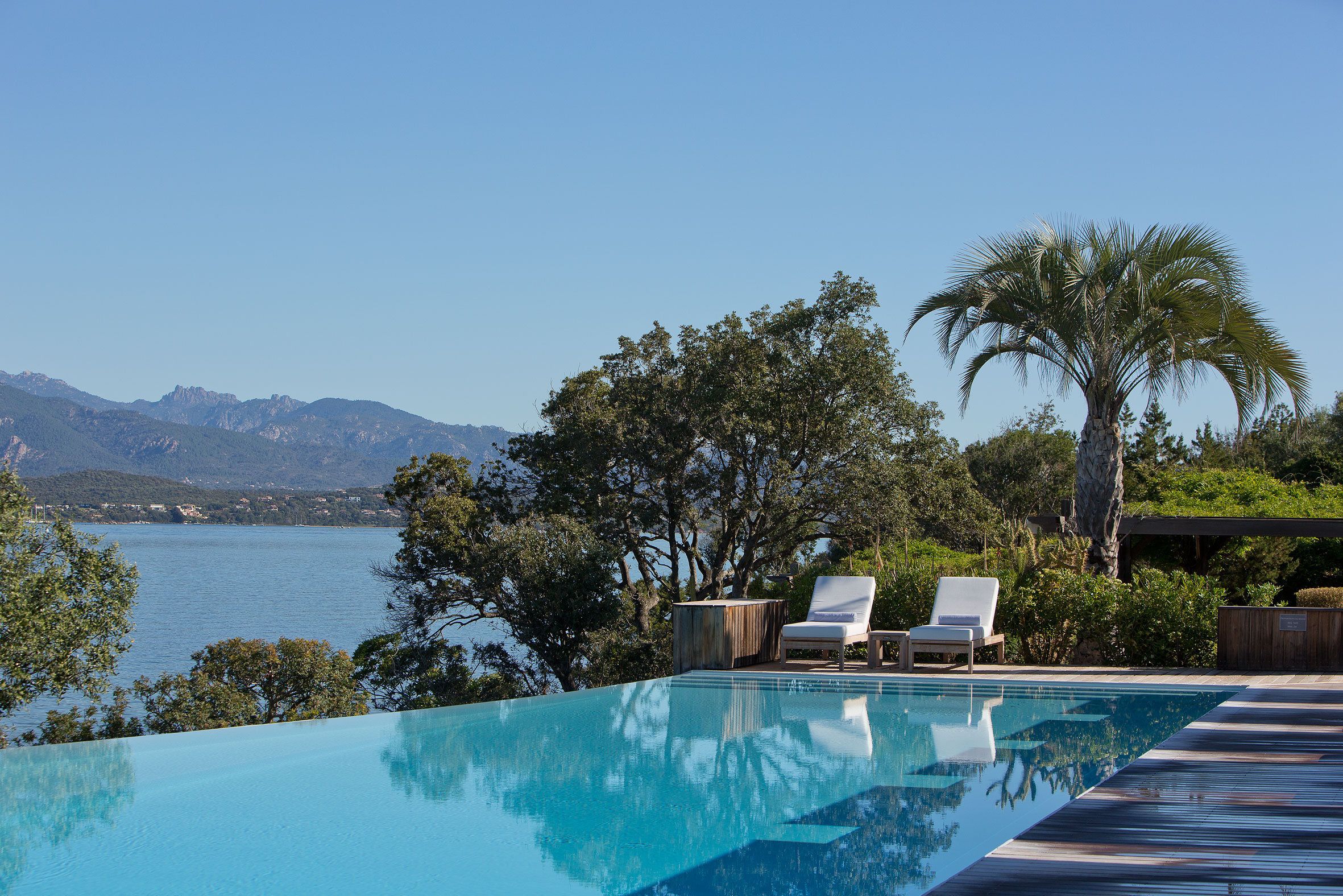 Una piscina a sfioro di un blu intenso, con due poltrone bianche, immersa nel verde e con vista su un lago e montagne.