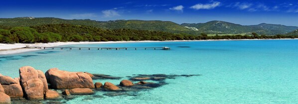 Une plage à l’eau turquoise et limpide, une jetée en bois et des affleurements rocheux.