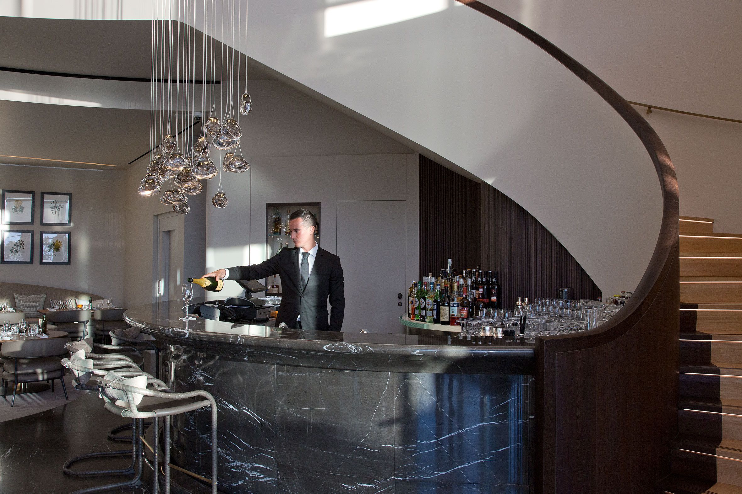 Zona de bar moderna con una barra de mármol curvada, taburetes y un bartender.