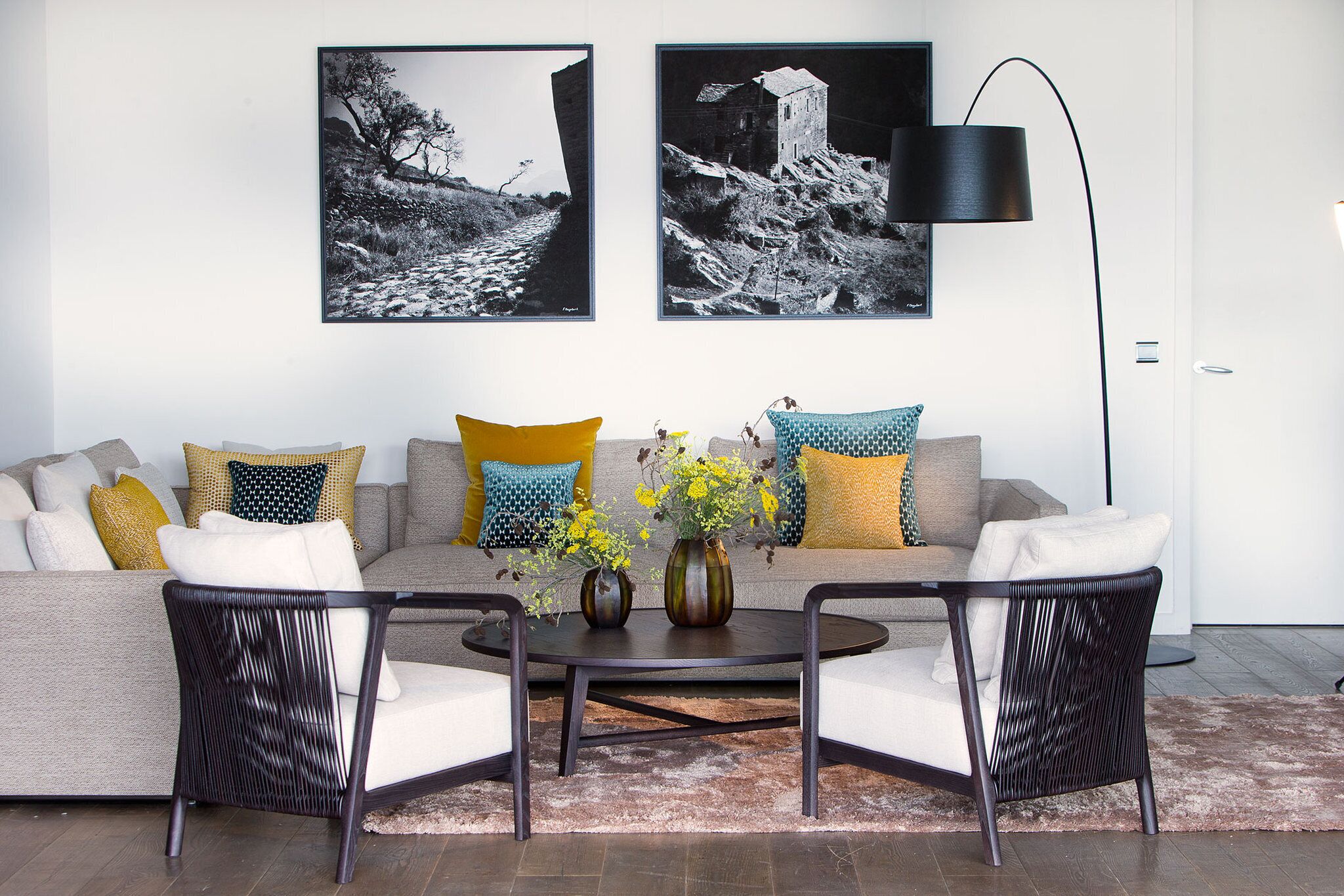 Ein modernes Wohnzimmer mit einer Couch, zwei Sesseln, einem Couchtisch und einer Stehlampe. An der Wand hängen zwei gerahmte Schwarz-Weiß-Fotos.
