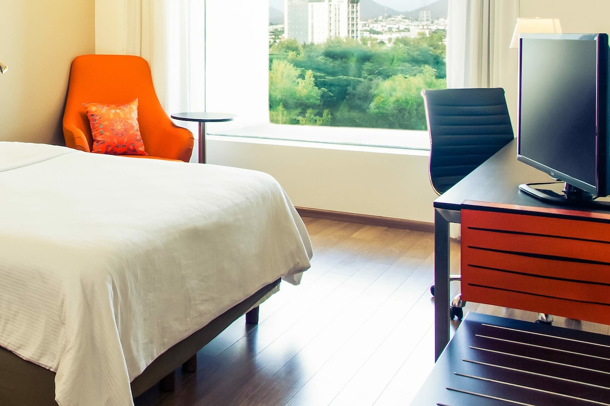 Habitación de hotel con una cama, un sillón naranja, un escritorio con una computadora y una ventana con vista a la ciudad.