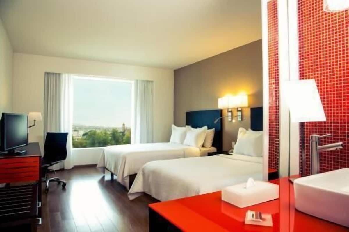 Habitación de hotel con dos camas, un escritorio con silla, un televisor y un baño con pared de azulejos rojos.