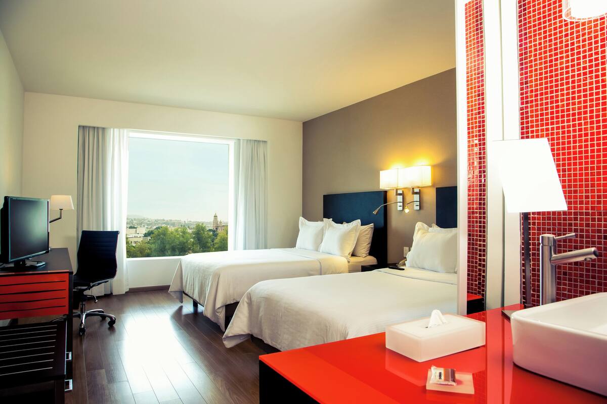Habitación de hotel con dos camas, un escritorio con computadora, una lámpara de escritorio, un ventanal grande con cortinas, una pared de acento roja y un baño con lavamanos y espejo.