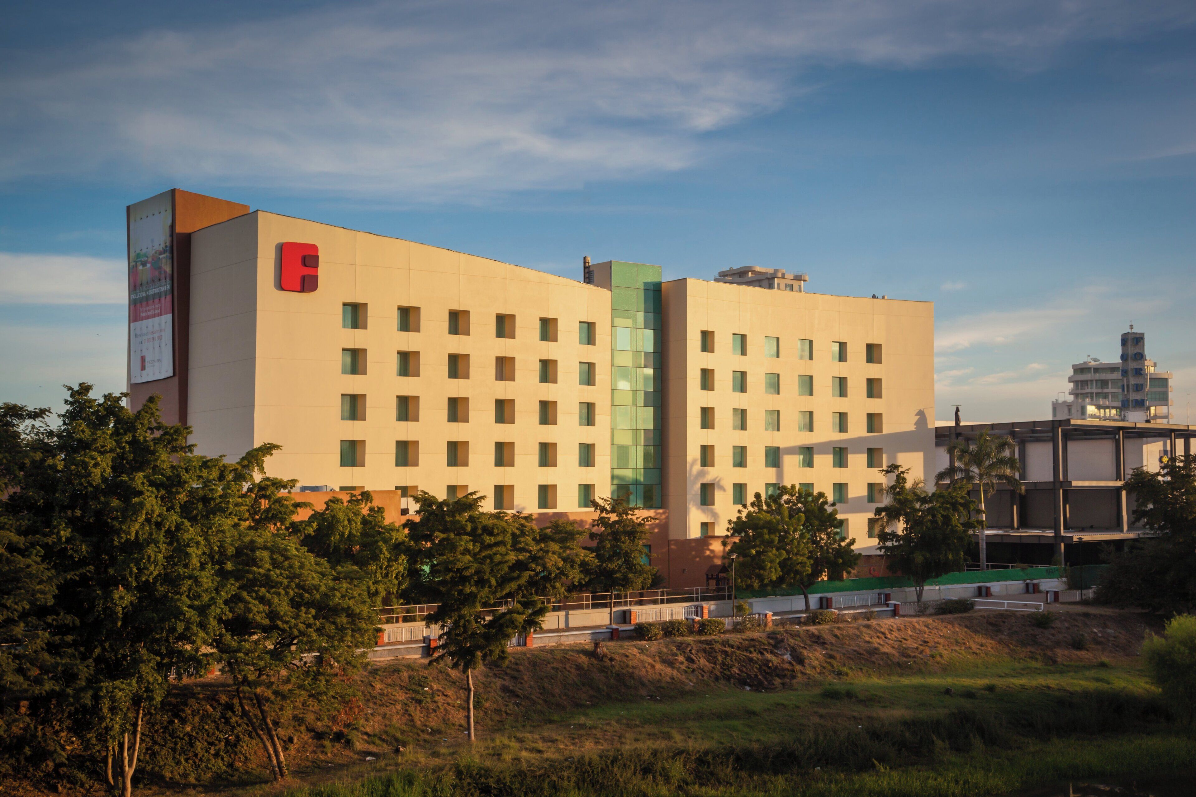 Un hotel de varios pisos con un logotipo rojo llamativo en la fachada, rodeado de vegetación y un cielo despejado.