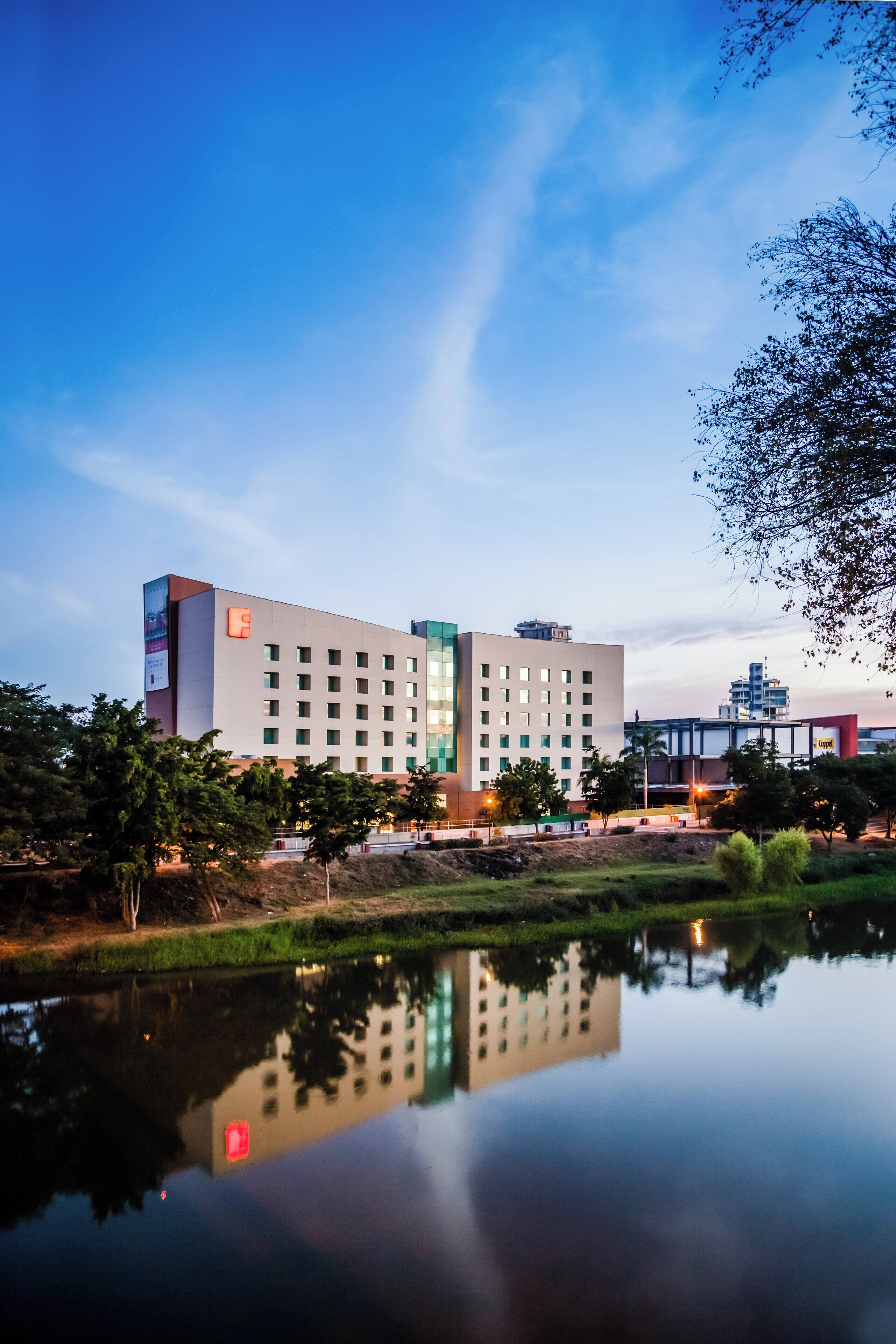 Un hotel moderno con una piscina reflectante en primer plano, rodeado de vegetación y un cielo despejado.
