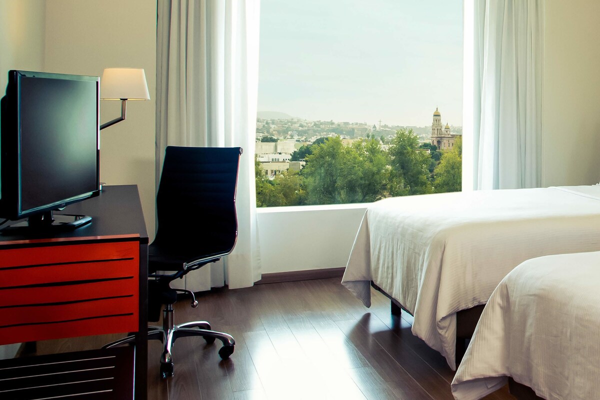 Habitación de hotel con cama, escritorio con computadora, silla y ventana que ofrece vista a la ciudad.