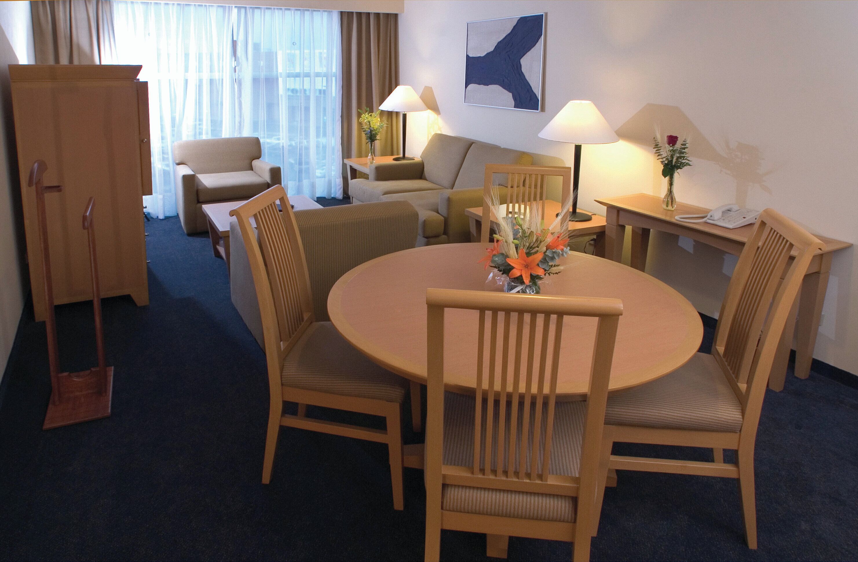 Une chambre d’hôtel avec une table de salle à manger ronde, quatre chaises en bois, un canapé, un bureau avec une lampe, un téléphone et un vase rempli de fleurs.