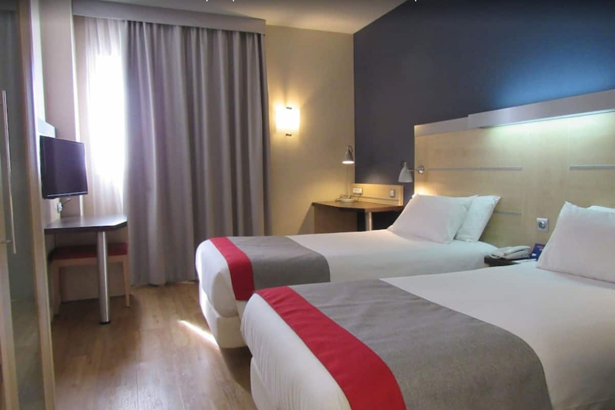 Ein Hotelzimmer mit zwei Betten, einem Schreibtisch, einem Fernseher und einem Fenster mit Vorhängen.