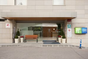 Entrada de hotel com um banco, plantas em vasos e placas indicando serviços como Holiday Inn Express e um balcão de atendimento Siemens.