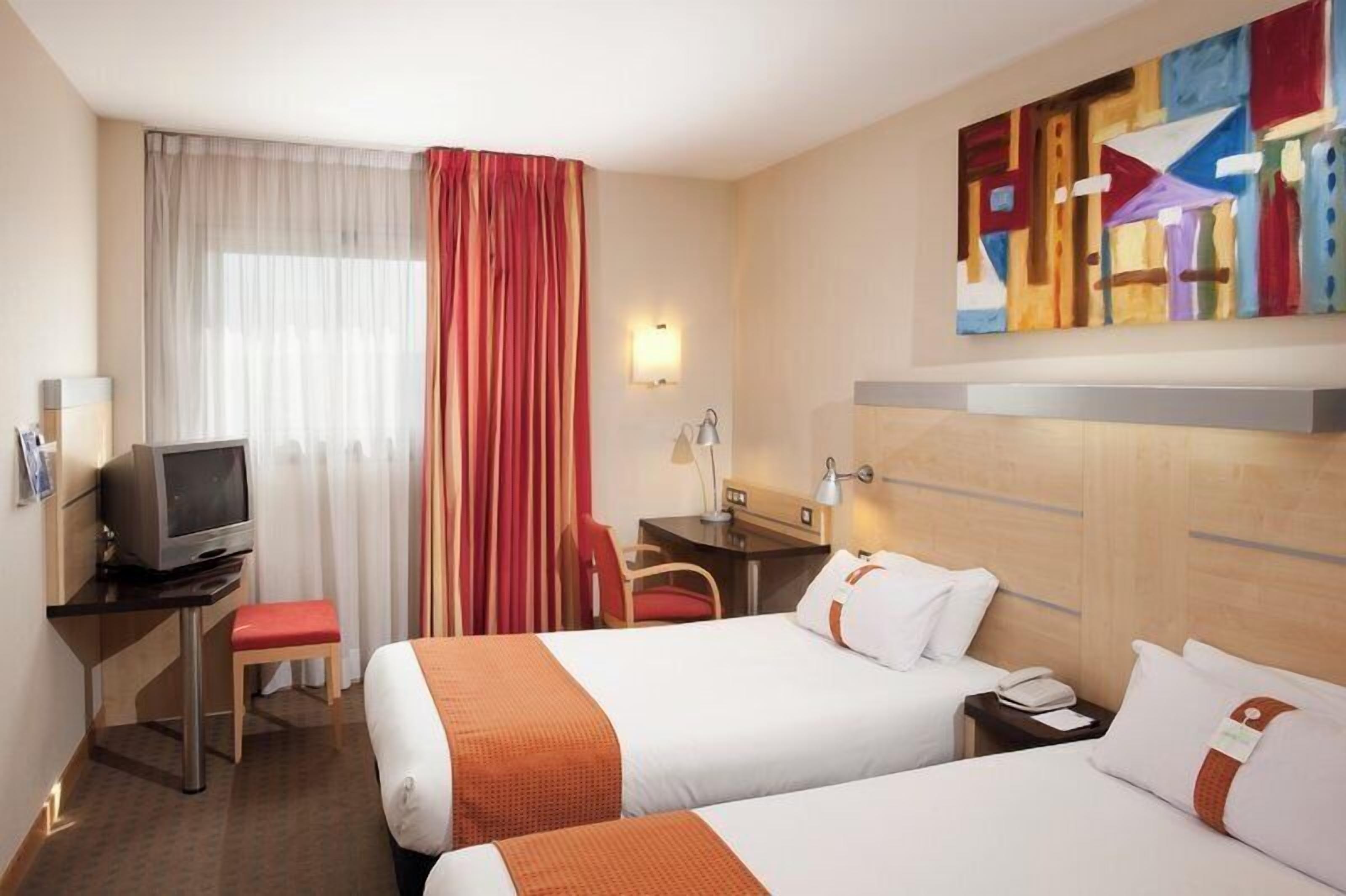 A hotel room with two beds, a desk with a television, a chair, a desk lamp, a phone, and a painting on the wall.