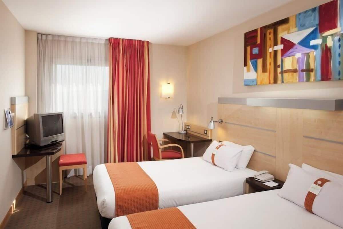 Ein Hotelzimmer mit zwei Betten, einem Schreibtisch mit Fernseher, einem Sessel, einer Schreibtischlampe, einem Telefon und einem Bild an der Wand.