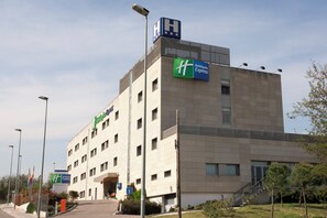 A multi-story Holiday Inn Express hotel with a prominent 'HH' sign on the roof, a 'T' parking sign, and a clear sky. - Holiday Inn Express Barcelona-Montmeló by IHG (Granollers)