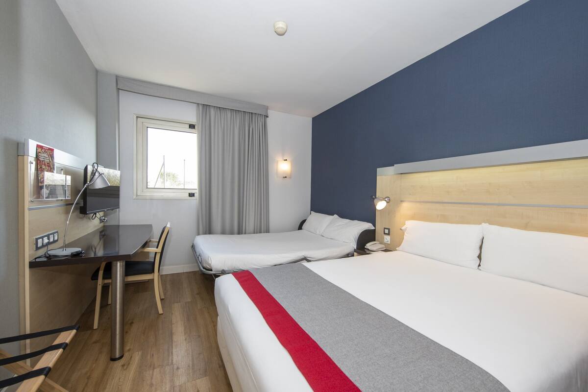 Ein Hotelzimmer mit Bett, Schreibtisch mit Stuhl, einem Fenster mit Vorhängen und einem an der Wand befestigten Telefon.