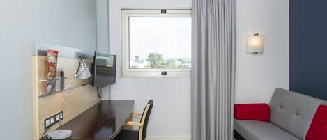 A modern hotel room with a wooden desk, a grey sofa, a television, and a window with curtains.