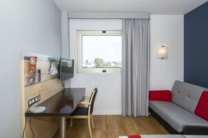 Una habitación de hotel moderna con un escritorio de madera, un sofá gris, un televisor y una ventana con cortinas.