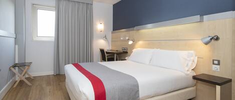 Una habitación de hotel con cama, escritorio, silla, lámpara y ventana con cortinas.