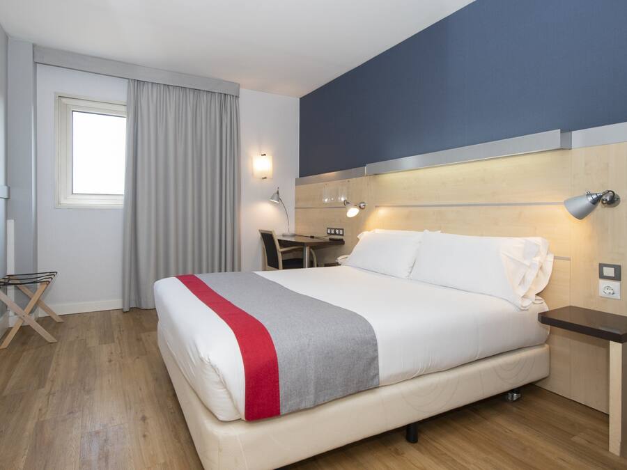 A hotel room with a bed, a desk, a chair, a lamp, and a window with curtains.