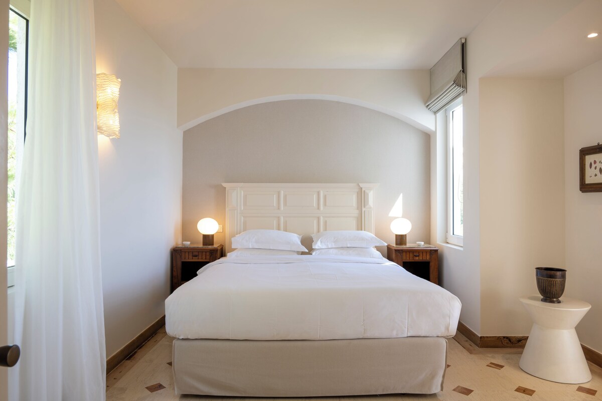 Une chambre avec un grand lit, deux tables de chevet avec des lampes, une petite table avec un bol, et une fenêtre avec des rideaux.