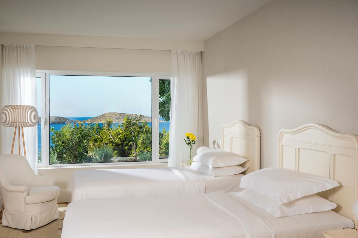 A bedroom with two beds, a chair, a lamp, and a view of the sea.