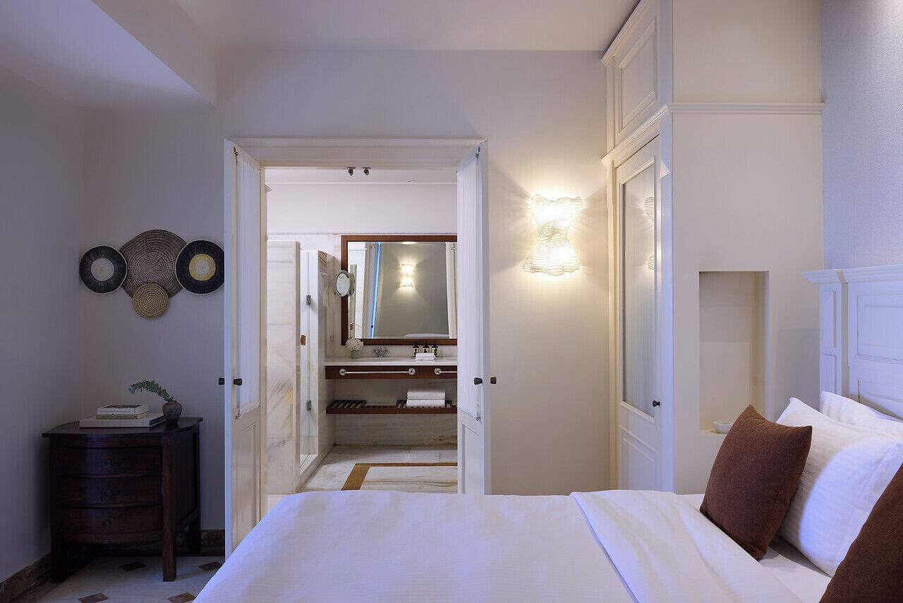 Ein Schlafzimmer mit einem Bett, einem Nachttisch, einer Kommode, einem Spiegel und einem durchgehenden Türspalt sichtbaren Badezimmer.