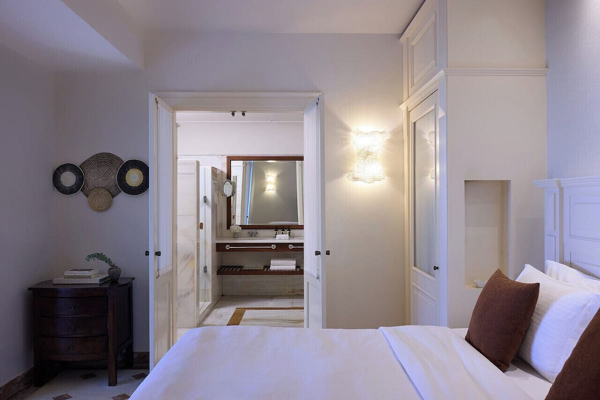 Un dormitorio con cama, mesita de noche, cómoda, espejo y baño visible a través de una puerta abierta.