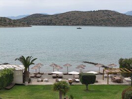 Vista costera con un barco en el agua, un patio con sombrillas y una montaña al fondo.