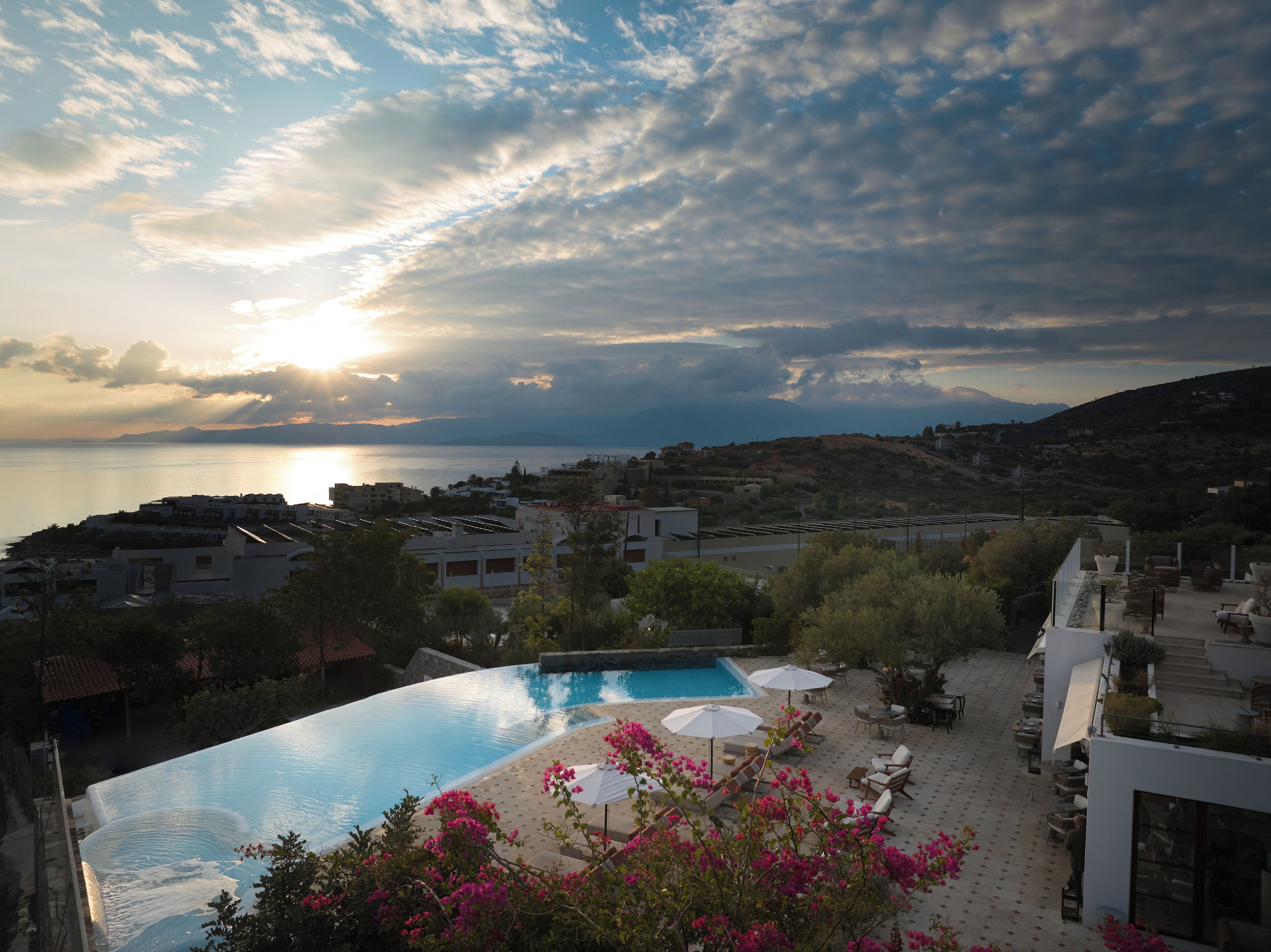 A swimming pool with lounge chairs and umbrellas, surrounded by greenery and buildings, with a view of the sea and mountains in the background.