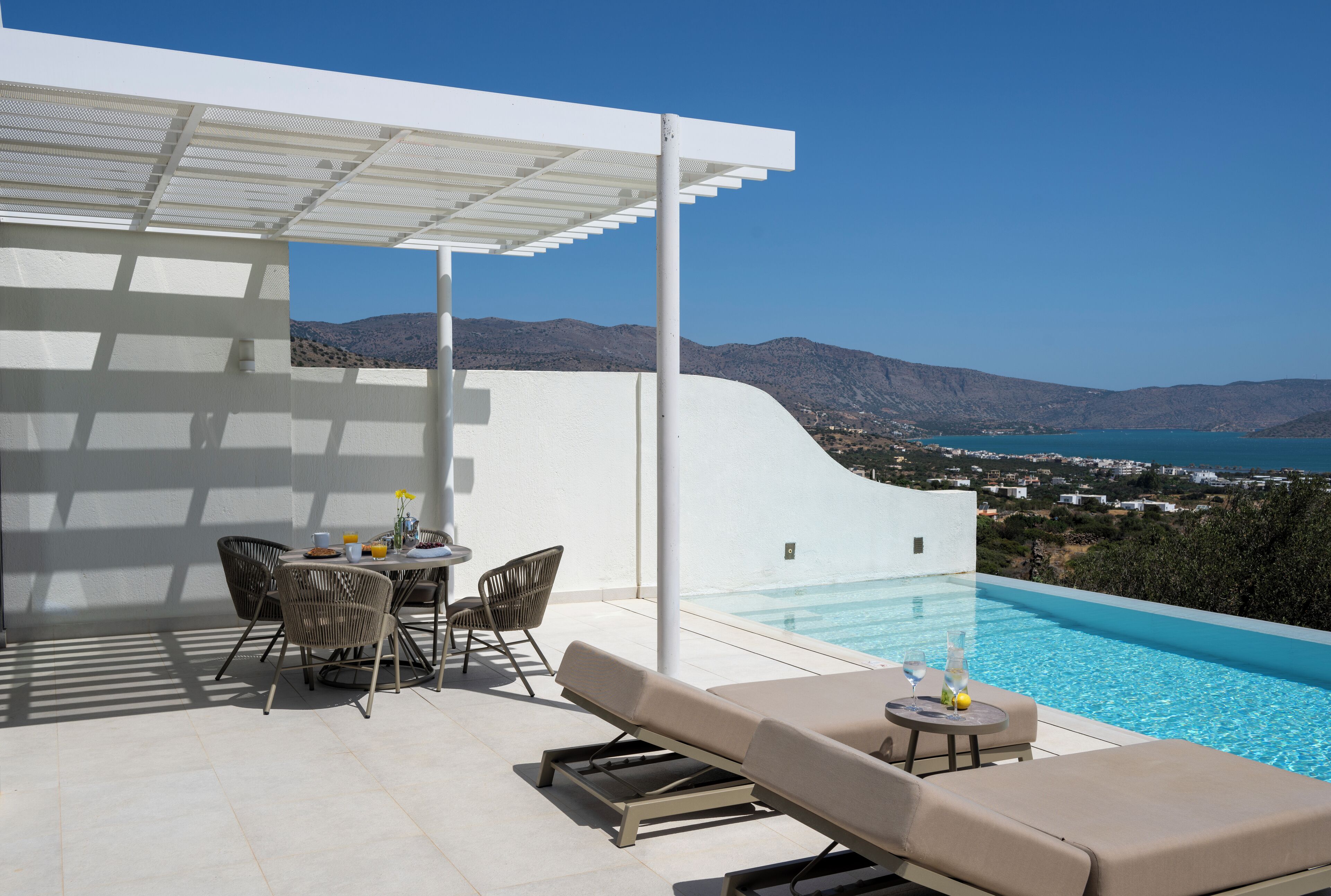 A modern outdoor pool area with a white wall, lounge chairs, and a dining table.