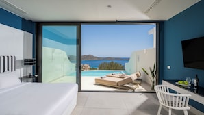 A modern hotel room with a large bed, a desk with a chair, a TV mounted on the wall, and a balcony with a pool and view of the sea.