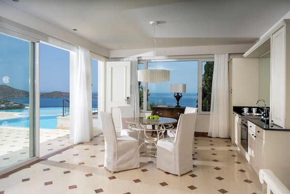 Amplia habitación con un gran ventanal con vistas a una piscina y al mar. Cuenta con una zona de comedor con mesa y sillas, una cocina con electrodomésticos modernos y una lámpara de araña grande.