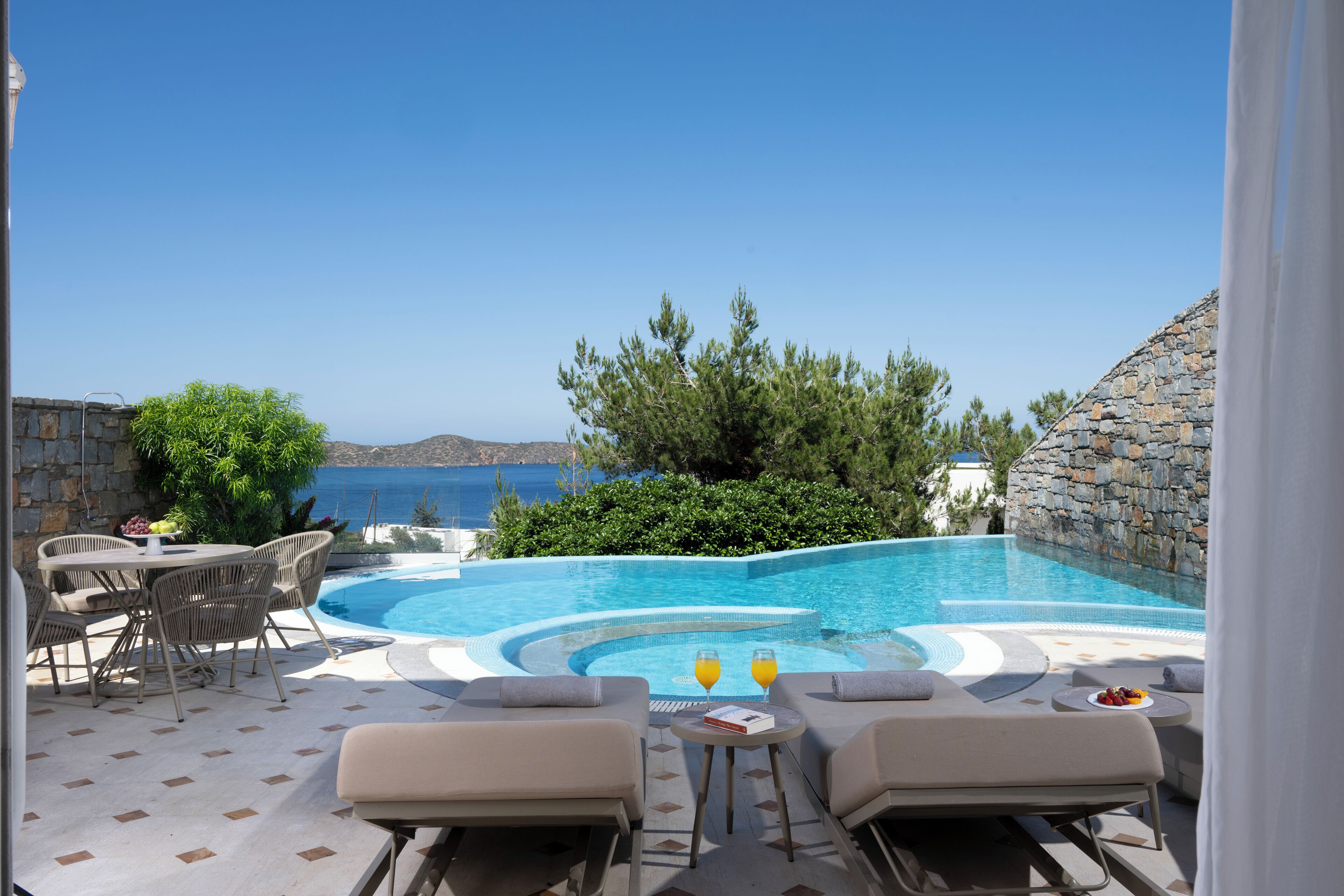 A pool area with lounge chairs and a table, surrounded by greenery and a stone wall.