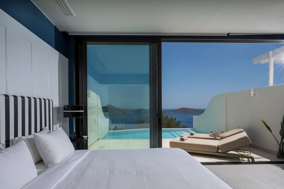 A modern hotel room with a large bed, a glass door leading to a balcony with a pool and a view of the sea.