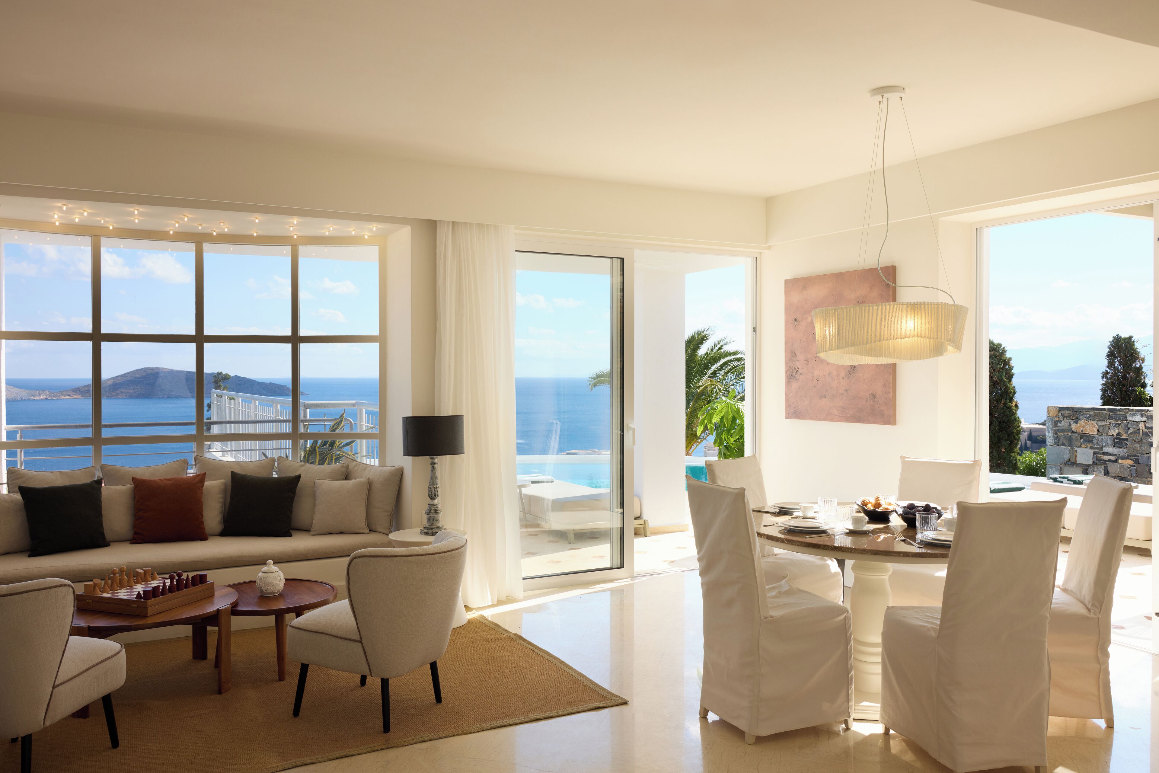 A spacious living room with a large window offering a view of the sea and mountains, a cozy sofa, a dining area with white chairs, and a chessboard.