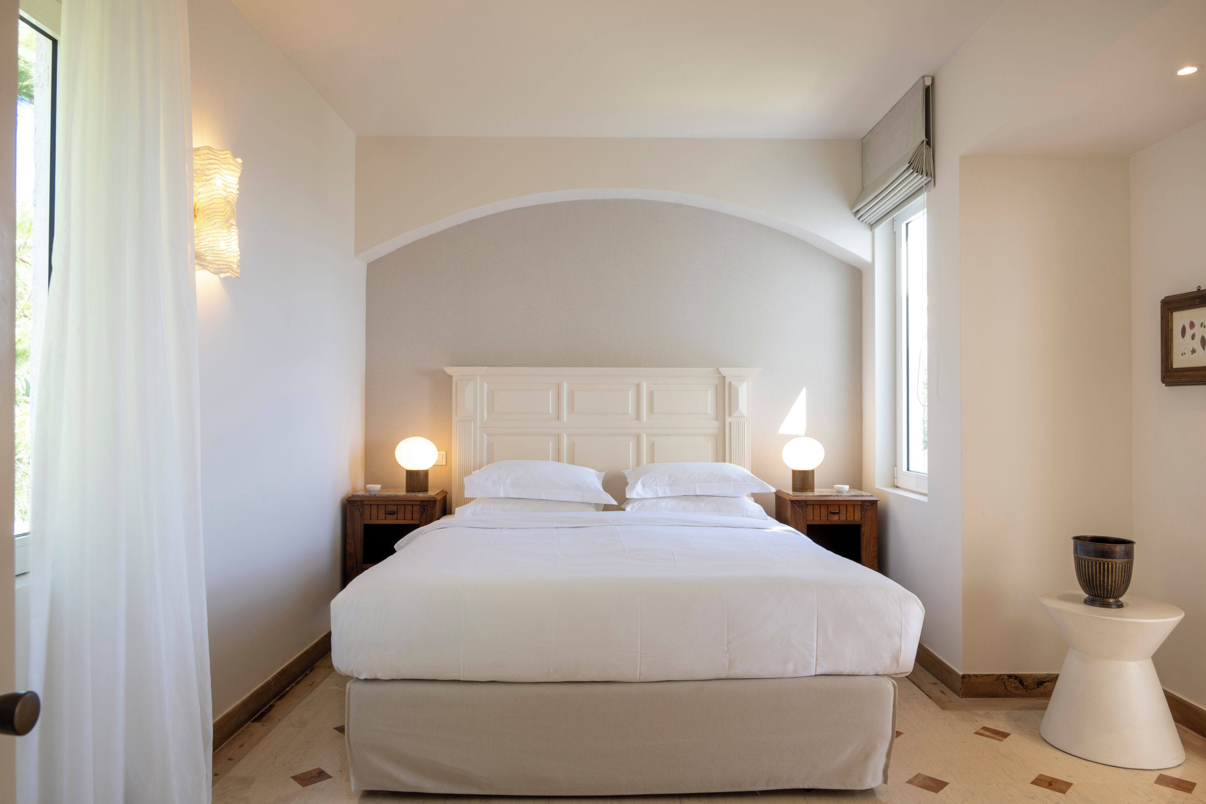Ein Schlafzimmer mit einem großen Bett, zwei Nachttischen mit Lampen, einem kleinen weißen Beistelltisch und einem Fenster mit Vorhängen.