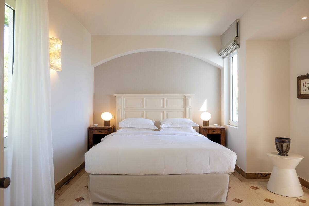 Un dormitorio con una cama grande, dos mesitas de noche con lámparas, una mesita pequeña con un cuenco y una ventana con cortinas.