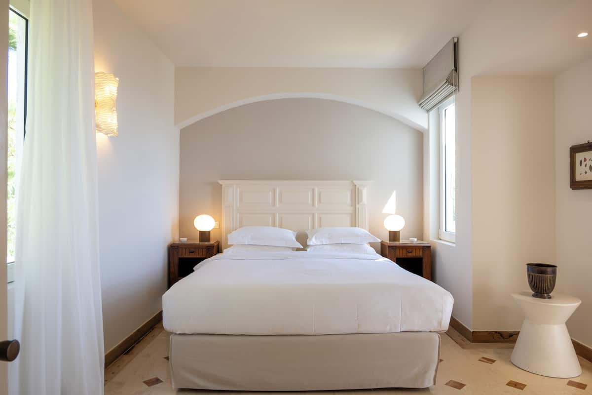 Ein Schlafzimmer mit einem großen Bett, zwei Nachttischen mit Lampen, einem kleinen weißen Beistelltisch und einem Fenster mit Vorhängen.