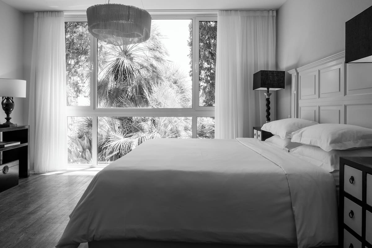 A monochrome hotel room with a large window, a bed with white linens, a bedside table with a lamp, and a dresser with drawers.