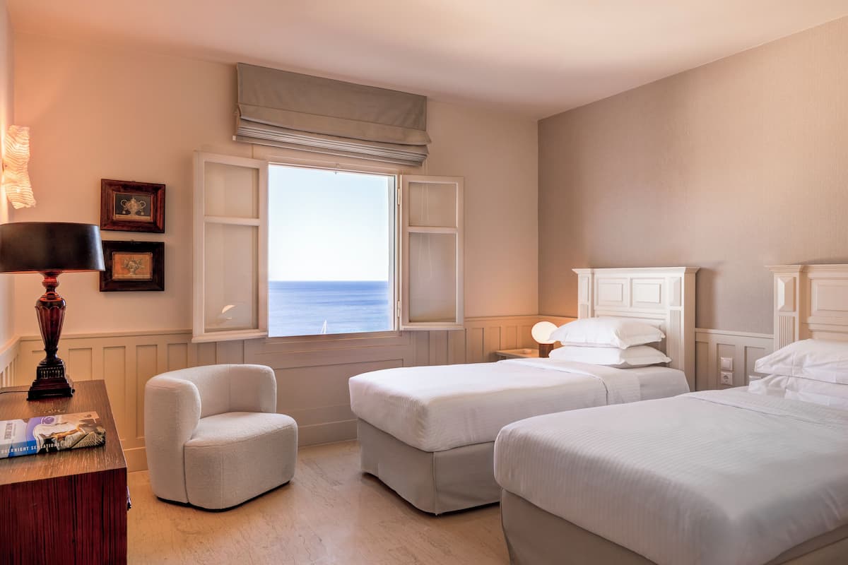 A hotel room with two beds, a chair, a desk, and a window with a view of the sea.