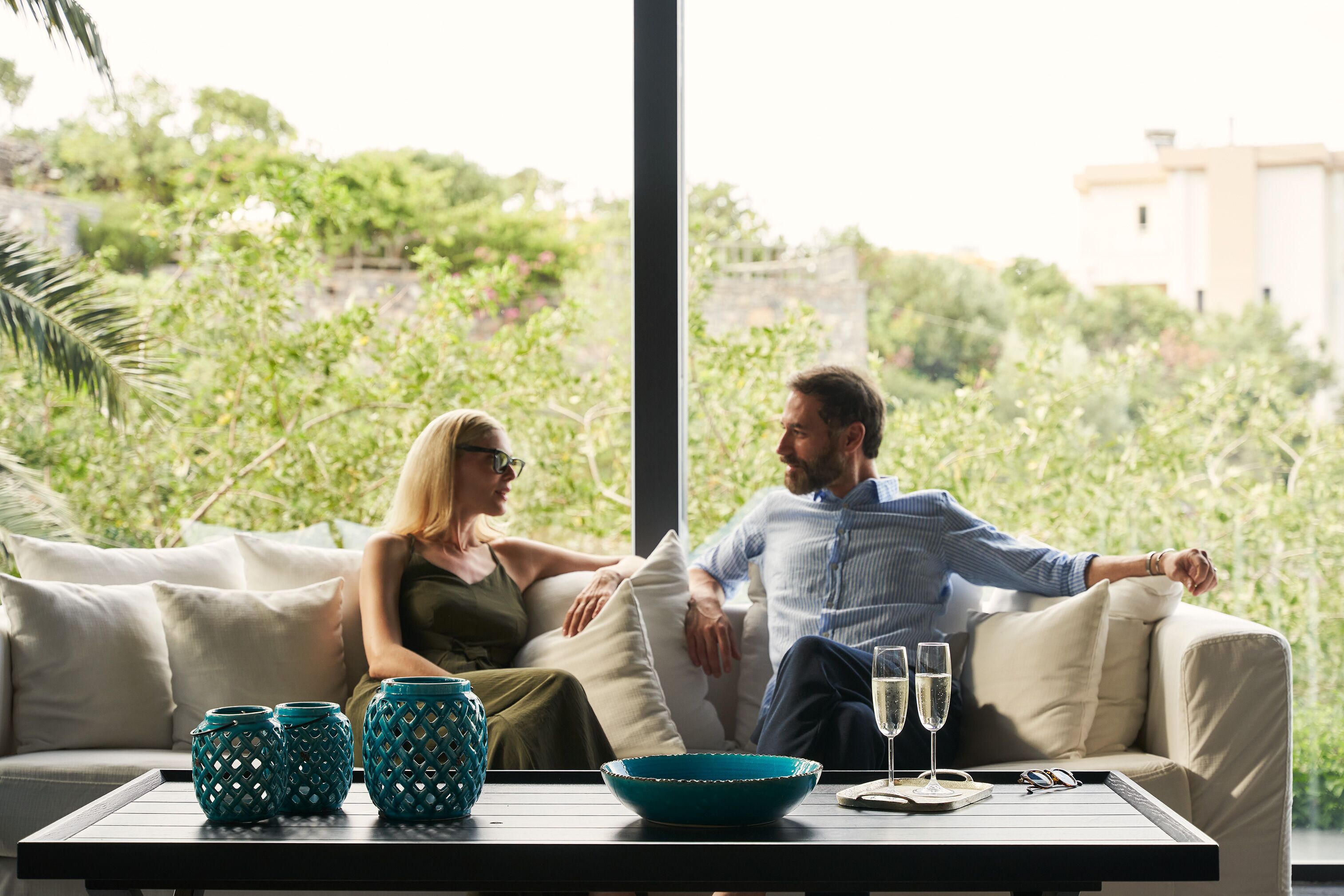 A couple on a beige sofa with a table in front of them, a window with a view of greenery, and two glasses of champagne.